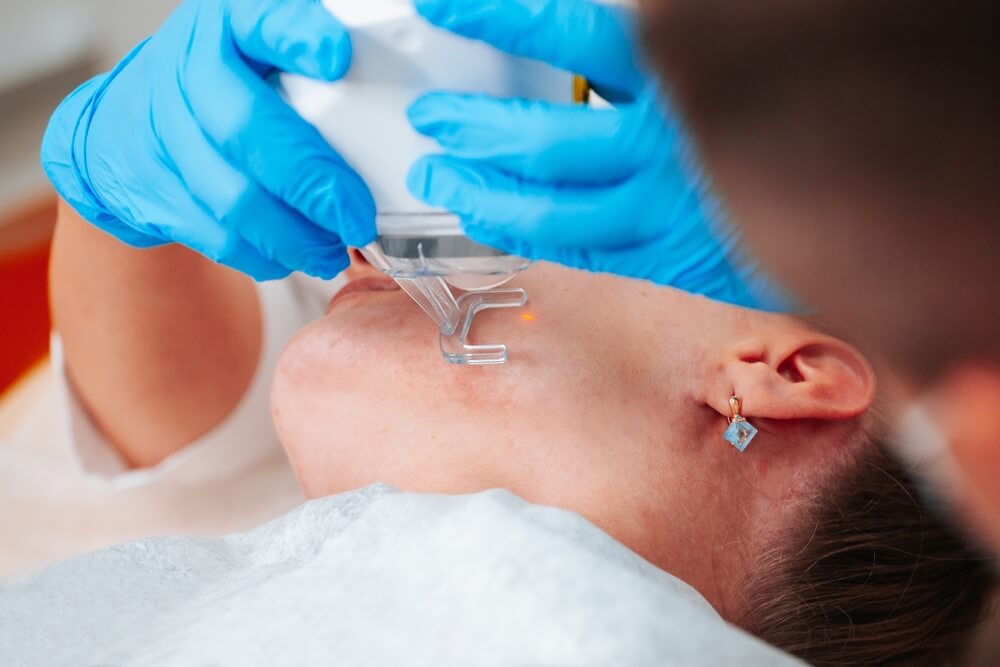 Lekarz w niebieskich rękawiczkach aplikuje żel lub krem na skórę szyi pacjenta podczas zabiegu dermatologicznego