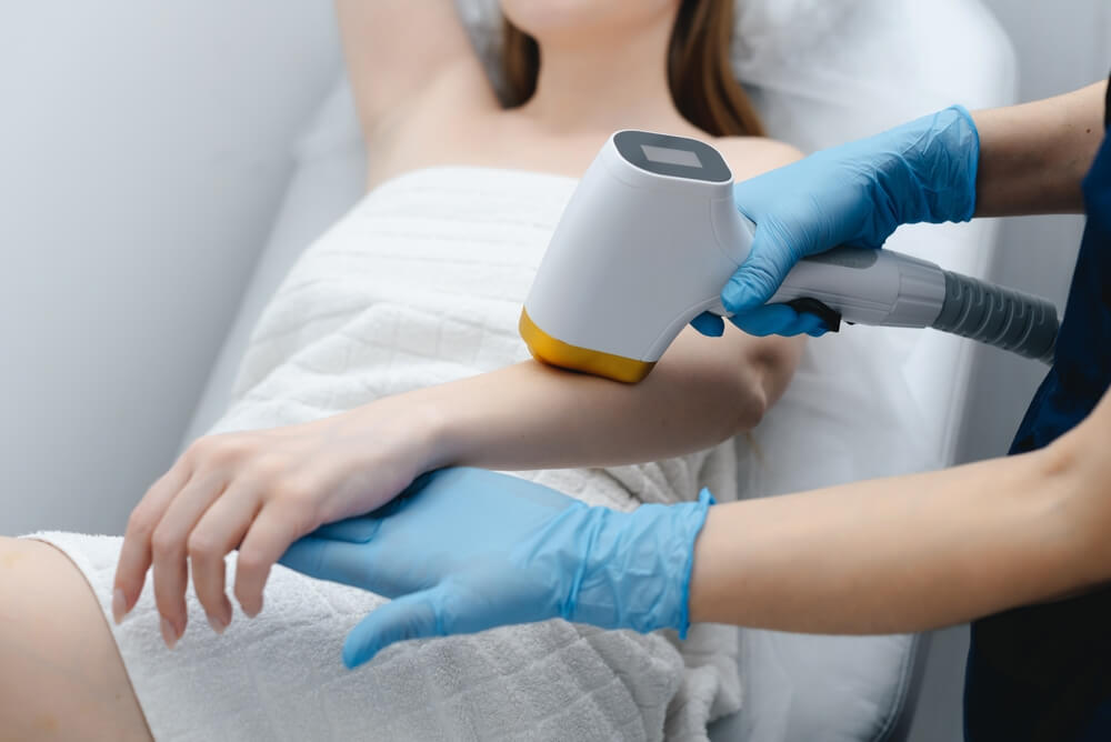 Dermatolog w niebieskich rękawiczkach wykonuje laserowe leczenie skóry na ramieniu pacjentki
